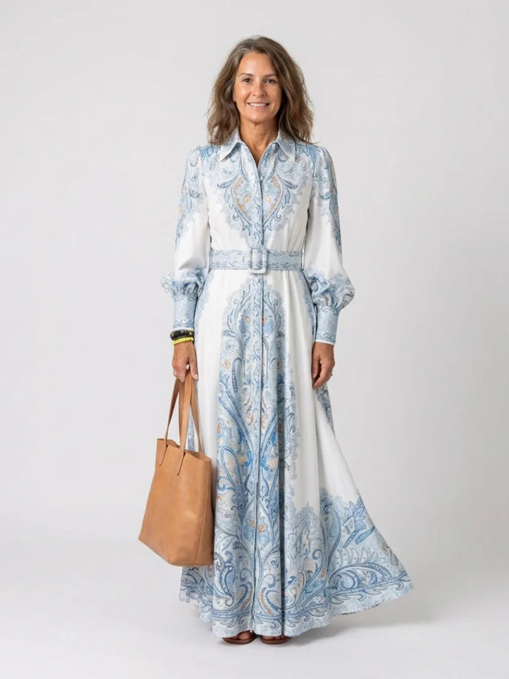 Woman wearing a high neck printed long sleeve maxi dress with paisley motif in soft blue tones and belted waist
